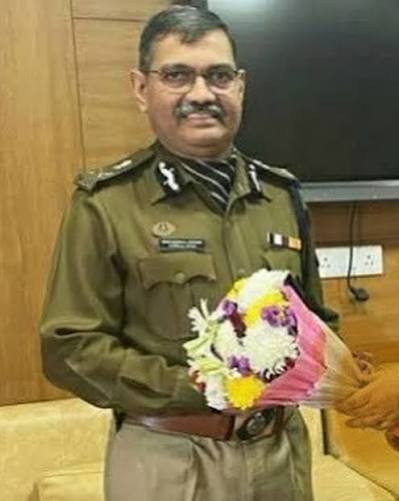 Narasimha Komar in Indian Police Service uniform, holding a bouquet of flowers presented to him indoors.