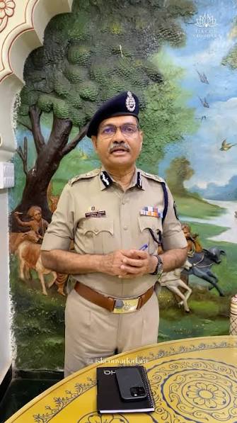 Narasimha Komar standing in uniform with a notebook, speaking, in front of a painted mural.