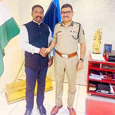 Narasimha Komar in uniform shaking hands with a visitor in his office, beside the Indian national flag.