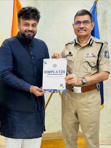 Narasimha Komar in uniform holding a printed tournament brochure alongside an organiser, with flags behind them.