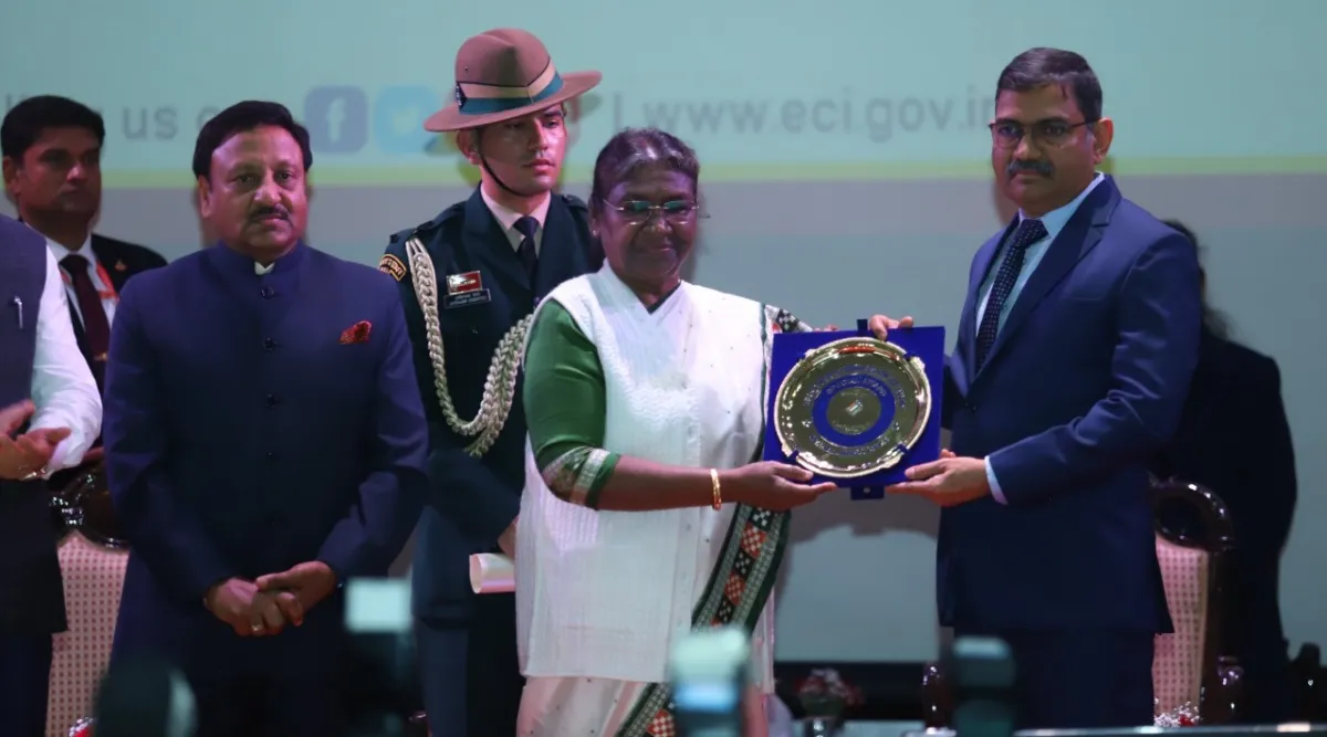 Narasimha Komar in a formal suit receiving a memento from the President of India on stage at an Election Commission of India ceremony.