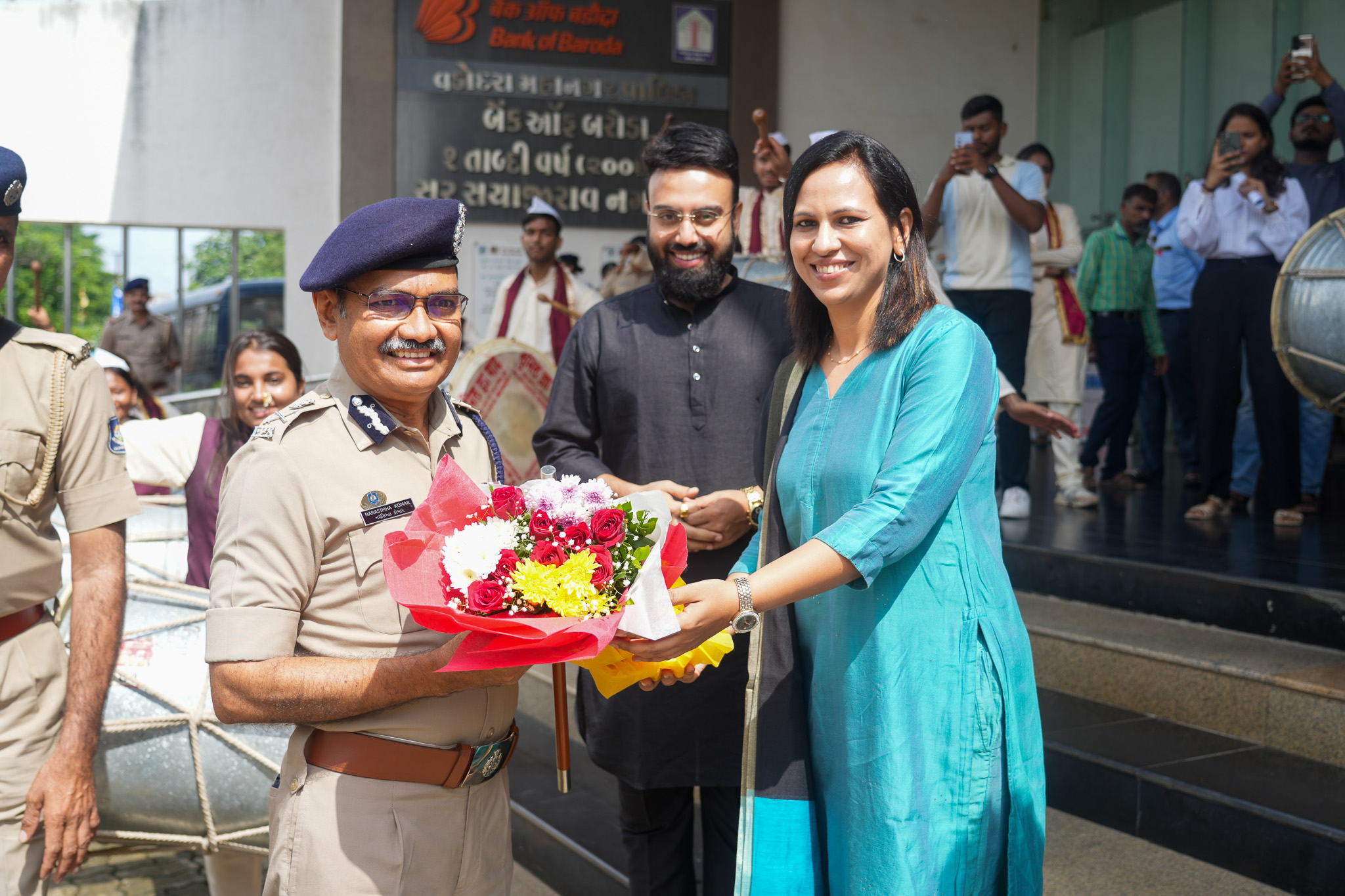 Narasimha Komar in ceremonial police uniform receiving a bouquet of flowers at a civic event, with a band and onlookers behind him.